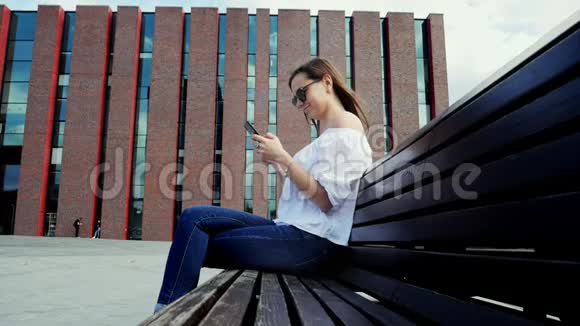 戴墨镜的快乐女人坐在街边的长凳上用手机现代的砖块建筑视频的预览图