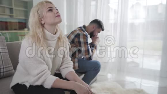 一个悲伤的金发白种人女人坐在沙发上的特写镜头她心烦意乱的男朋友或丈夫坐在后台视频的预览图