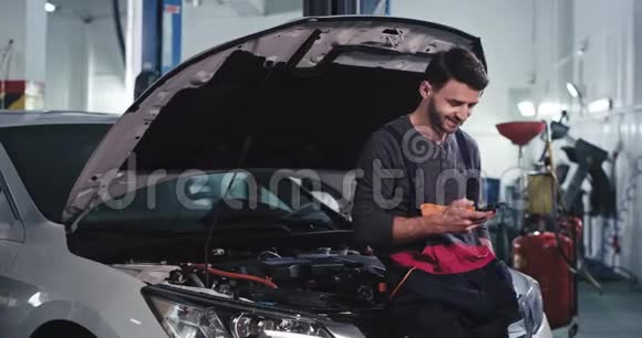 一个穿制服的工人微笑着在汽车修理厂修理汽车时看了看手机视频的预览图