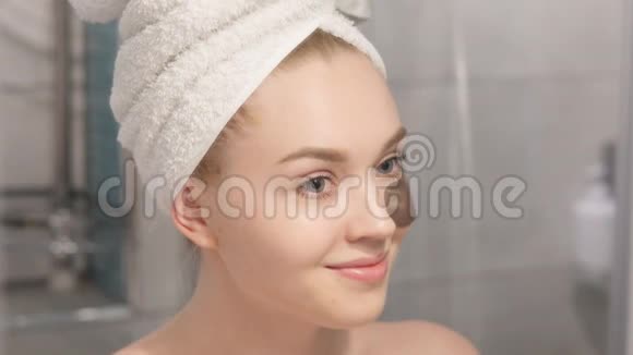 一张美丽的年轻女子的肖像穿着浴袍和毛巾站在浴室里在她的眼睛下敲击着补水补丁视频的预览图