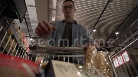 一个戴眼镜的白人年轻人把东西放在购物车里穿过一个大杂货店的货架视频的预览图