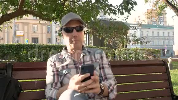 白种人坐在公园里的长椅上点击智能手机喝水视频的预览图