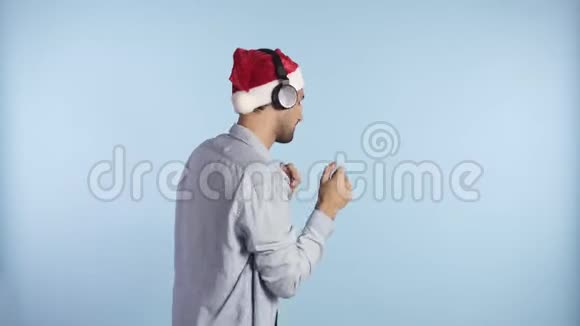 戴着圣诞红帽子戴着耳机听着他最喜欢的音乐在无忧无虑的环境中跳舞的英俊男子视频的预览图