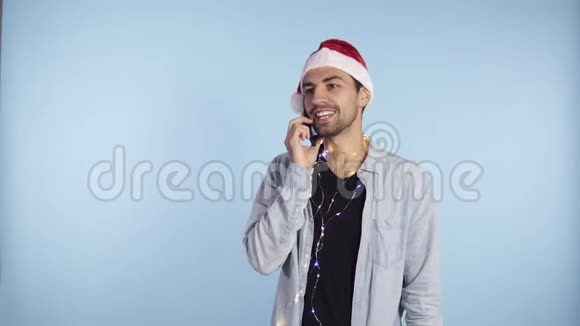 年轻快乐的情感男人戴着圣诞圣诞老人帽脖子上戴着花环站着用手机说话男子视频的预览图