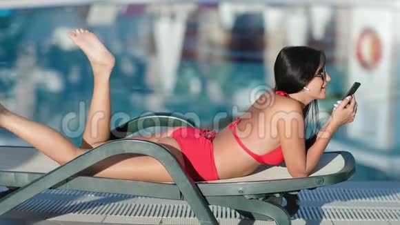 穿着泳衣的时尚女士在躺椅上放松用智能手机听音乐视频的预览图