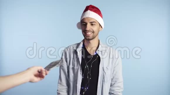 年轻的快乐男人收到纸币美元钞票戴着圣诞帽站在蓝墙边数钱视频的预览图