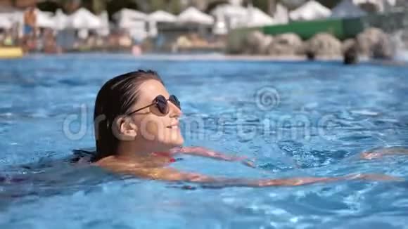 戴墨镜的微笑女子漂浮在游泳池周围是蓝色的波纹特写视频的预览图