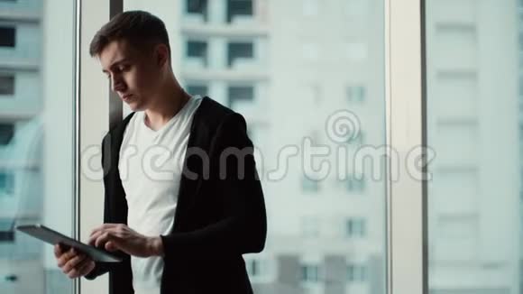 英俊自信的年轻人在现代办公室的一个大窗户附近使用平板电脑视频的预览图