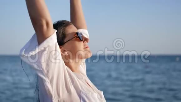 穿着白色衬衫的可爱的晒黑轻松的女孩举起手在海上享受自由视频的预览图