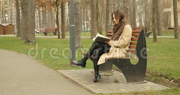 女人在长凳上读完一本书站起来戴着棕色的麦金托什和带帽子的围巾离开秋天的公园视频的预览图