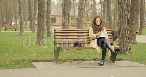 年轻迷人的女性独自坐在公园的长凳上读着一本穿着棕色战壕和围巾的书学习概念视频的预览图
