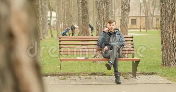 年轻的胡须男拨打电话开始在电话上交谈微笑着享受着秋公园坐在长凳上的谈话视频的预览图