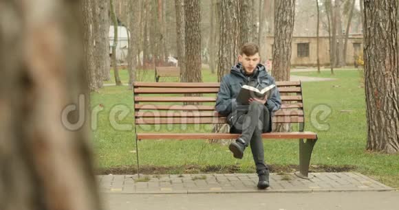 秋天公园里一个长胡子的年轻人穿着蓝色夹克坐在长凳上看书视频的预览图