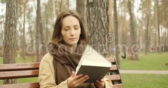 迷人的女人打开一本书翻开白纸然后开始阅读一本小说而独自坐在秋天寒冷的公园里穿着衣服视频的预览图