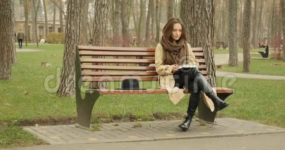 可爱的女性开始阅读一本书翻书页而坐在秋天公园的长凳上独自穿着战壕和棕色围巾视频的预览图