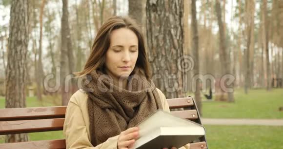 可爱专注的女人翻书页独自坐在寒冷的秋天公园的长凳上戴着棕色的围巾看书视频的预览图