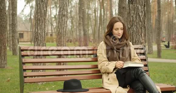 年轻可爱的女孩坐在公园里的长凳上读着一本放在她附近的黑帽子的书阅读概念视频的预览图