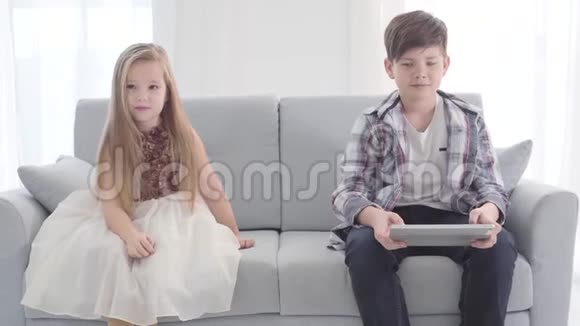 两个高加索儿童坐在沙发上的肖像在平板电脑屏幕上看东西的男孩穿着漂亮衣服的女孩视频的预览图