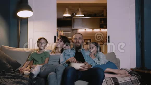 快乐微笑的年轻白种人家庭与两个孩子在家看电影沙发一起笑娱乐概念视频的预览图