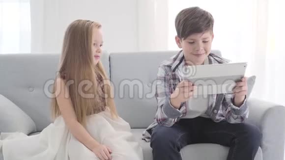 迷人的白人女孩坐在沙发上男孩拿着平板电脑害羞漂亮的小女人看着她的爱照相机视频的预览图