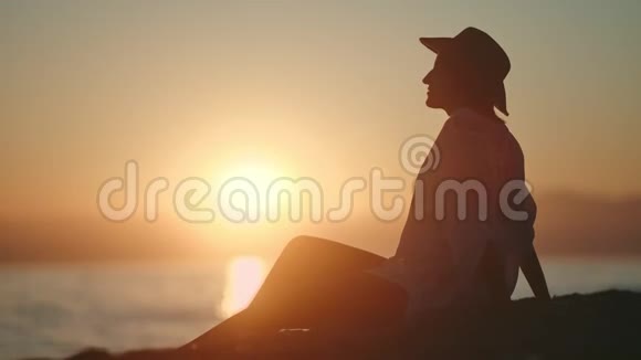 放松的女游客戴着帽子的剪影在海岸边欣赏着令人惊叹的自然日落视频的预览图