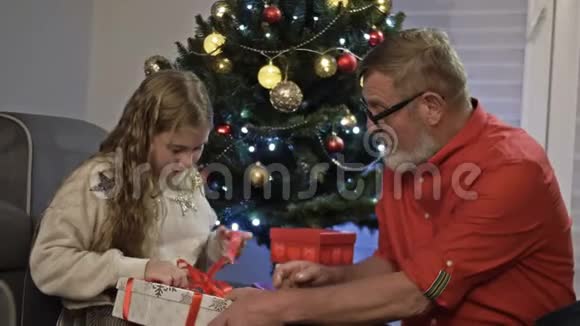 祖父和孙女靠近圣诞树女孩打开礼物高兴起来新年快乐视频的预览图