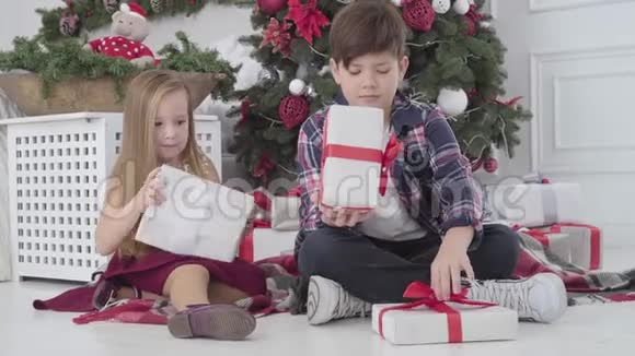 漂亮的白人女孩和男孩把礼物放在圣诞树下室内孩子们回头看装饰的杉树视频的预览图