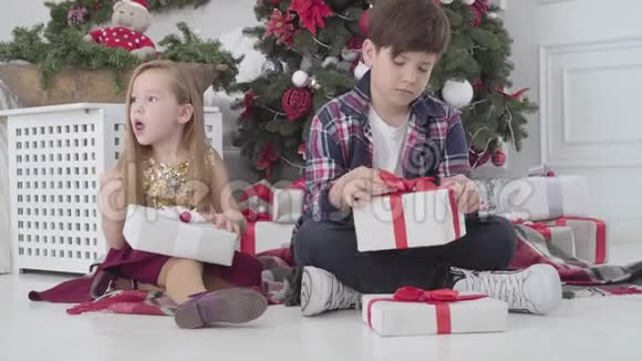 可爱的高加索儿童坐在圣诞树下带着礼品盒环顾四周的肖像女孩和男孩视频的预览图