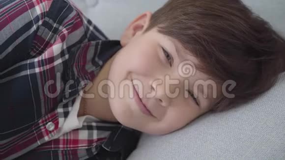 白人男孩的特写棕色头发和灰色的眼睛看着相机和微笑快乐的孩子躺在软榻上视频的预览图