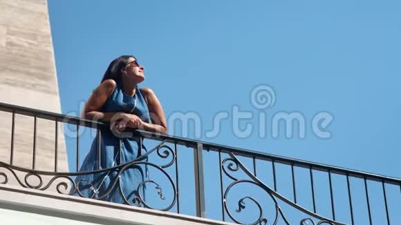 穿着裙子站在阳台上享受阳光的女性放松的优雅晒黑视频的预览图