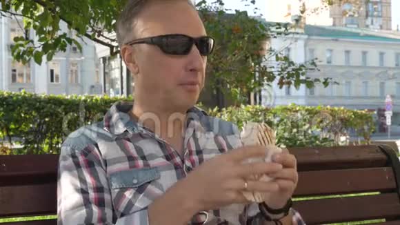 戴墨镜的白人中年男子在长凳上吃大三明治视频的预览图