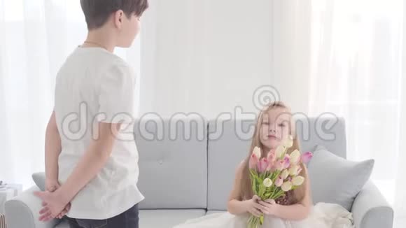 漂亮的白人孩子穿着裙子坐在沙发上捧着一束郁金香抬头看着男孩恋爱中的儿童视频的预览图