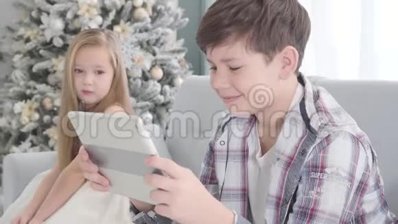 可爱的高加索女孩坐在背景上看着男孩玩平板电脑的肖像害羞的小姑娘视频的预览图