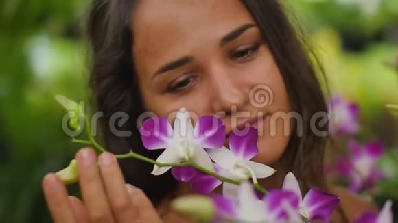 在花园里微笑着美丽的年轻女子一个女人摸着兰花享受它的气味慢动作视频的预览图