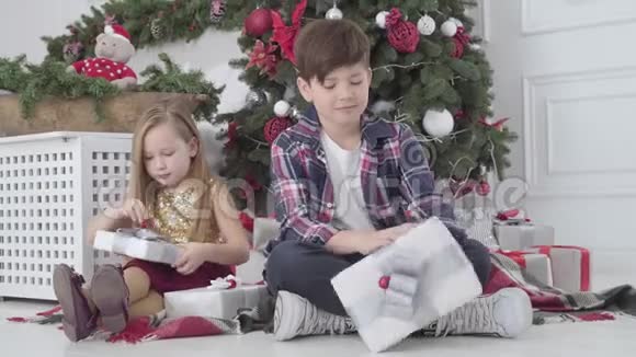 小男孩和小女孩拿着礼物坐在新年树下圣诞前夜快乐儿童的肖像假期视频的预览图