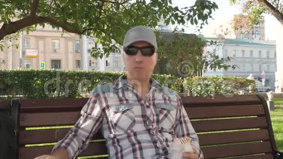 戴墨镜的白种人坐在公园的长椅上吃着智能手机的点击视频的预览图