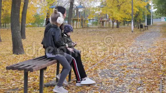 一位年轻的母亲两个孩子坐在秋天公园的长凳上友好的家庭观念视频的预览图