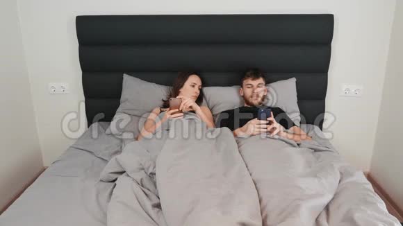 已婚夫妇用智能手机躺在床上一对夫妇躺在床上看着智能手机社会Y的交流视频的预览图
