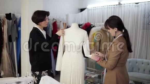 两名白种人的女裁缝在白色婚纱上合作并将未完成的服装钉在时装模特身上视频的预览图
