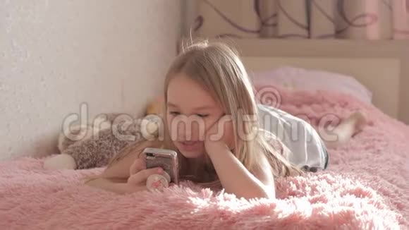 可爱的女孩在房间里躺在床上玩智能手机视频的预览图