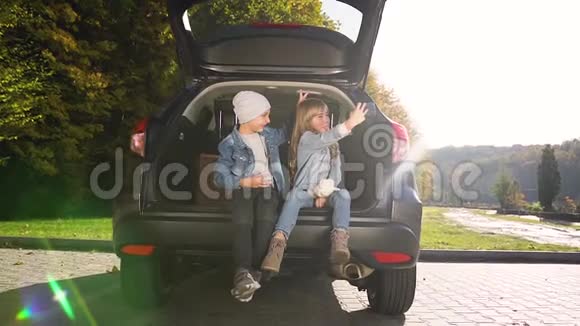 满意的快乐少年男孩和女孩穿着牛仔裤的衣服坐在后备箱里在女孩的手机上做有趣自拍视频的预览图