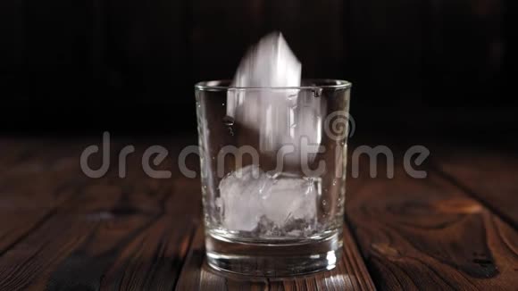 在黑暗的木制背景上冰块落入空玻璃中视频的预览图