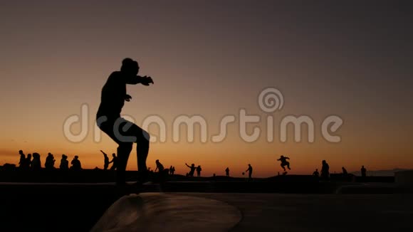 少年跳滑板的剪影骑长板夏日夕阳背景洛斯威尼斯海滨溜冰场视频的预览图