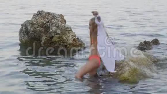 穿着泳衣和长袍的年轻女孩在夏天海滩的海水中的石头之间行走海水中的少女视频的预览图