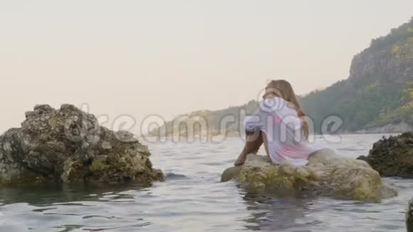 体贴的少女穿着白色湿衣坐在海上的石头上的山景坐在岩石上的旅游女孩视频的预览图