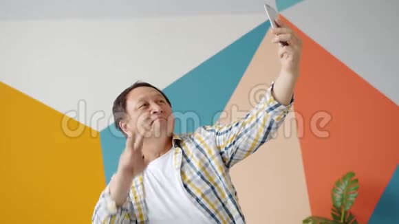 欢乐的亚洲男人在家里用智能手机拍照显示手势视频的预览图