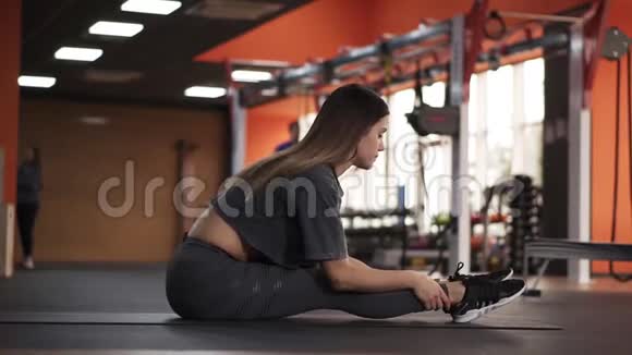年轻漂亮的瑜伽女人坐在垫子上的地板上伸展身体向腿倾斜年轻女孩做瑜伽视频的预览图