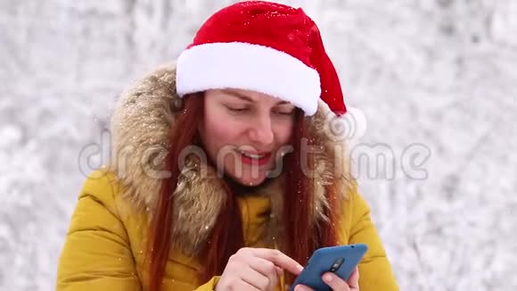 穿着温暖的冬衣和圣诞帽的年轻女孩使用智能手机体验快乐的情绪聊天赢概念视频的预览图