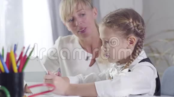 旁边看着可爱的白种人女学生坐在桌子旁拿着平板电脑当中年家教解释着她在视频的预览图