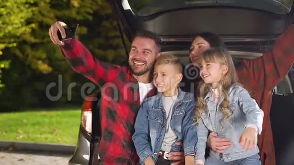 现代美丽的微笑家庭的迷人肖像坐在汽车后备箱里在爸爸的身上拍着有趣照片视频的预览图